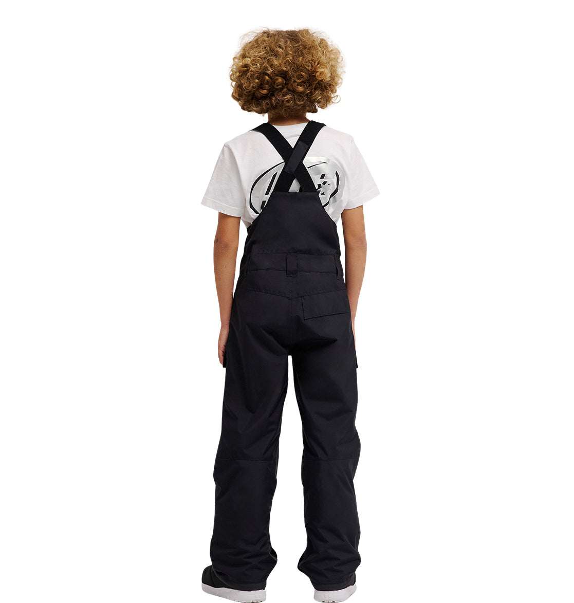 A child with curly hair, wearing the DC Shoes Roadblock Youth Bib over black insulated bib overalls and a white graphic T-shirt, stands facing away from the camera against a plain white background.