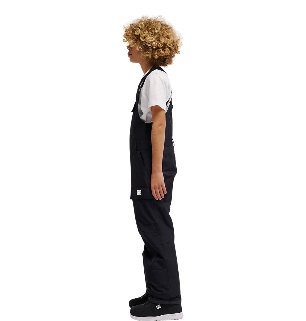 A child with curly blond hair stands in profile against a plain white background, wearing a white T-shirt, black sneakers, and the DC Shoes Roadblock Youth Bib—sleek black insulated overalls.