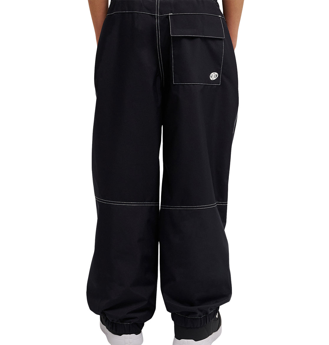 A person is shown from the waist down, wearing black DC Shoes Trackstar Baggy Youth Pants featuring white stitching, elastic cuffs, and a large back pocket. These cargo pants are designed for enhanced snow performance and are set against a white background.