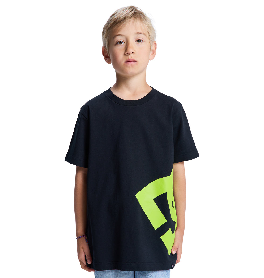 A young boy with short blonde hair faces forward, wearing the Reebok Lanai Youth Hss black t-shirt featuring a large yellow-green side graphic, paired with light blue jeans. The background is plain white.