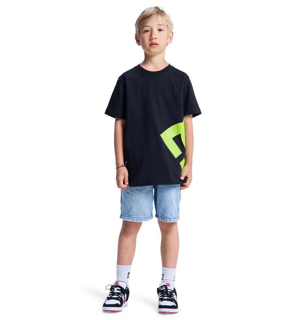 A young boy stands facing forward against a white background, wearing the Reebok Lanai Youth Hss black T-shirt with a neon green geometric design, light blue denim shorts, white socks, and black and white sneakers.