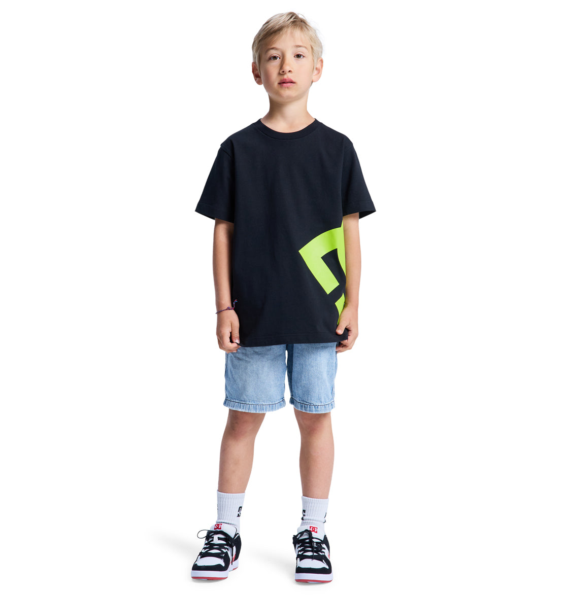 A young boy stands facing forward against a white background, wearing the Reebok Lanai Youth Hss black T-shirt with a neon green geometric design, light blue denim shorts, white socks, and black and white sneakers.