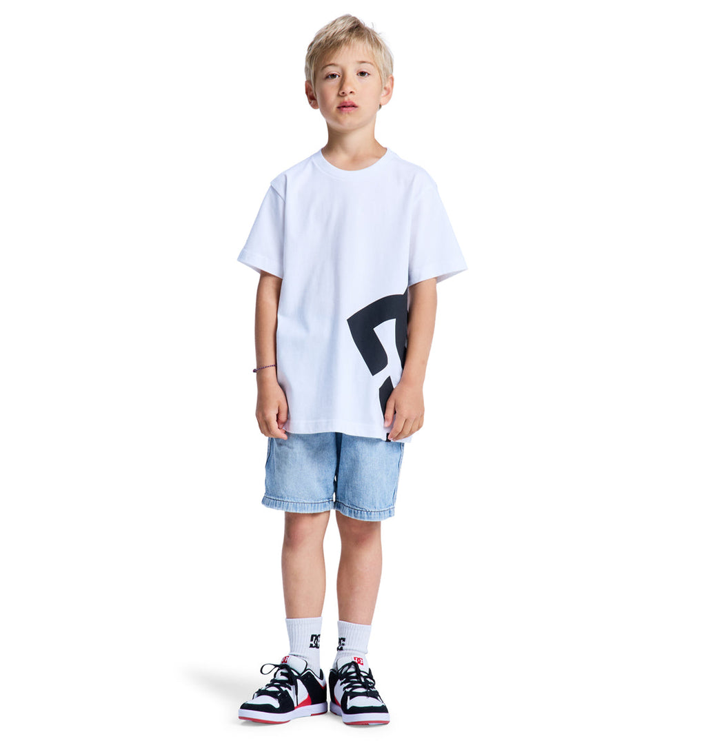 A young boy stands against a plain white background wearing the Reebok Lanai Youth Hss T-shirt, denim shorts, white socks, and black-and-white sneakers.