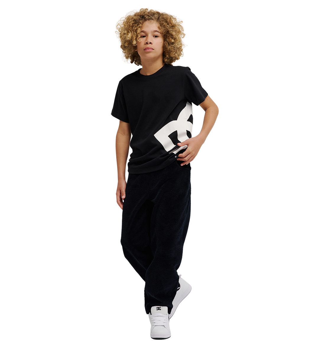 A young person with curly blond hair wears the DC Shoes DC Lanai Tee Youth, a black relaxed-fit graphic skate shirt with a bold white logo, along with loose black pants and white sneakers, standing against a plain white background.