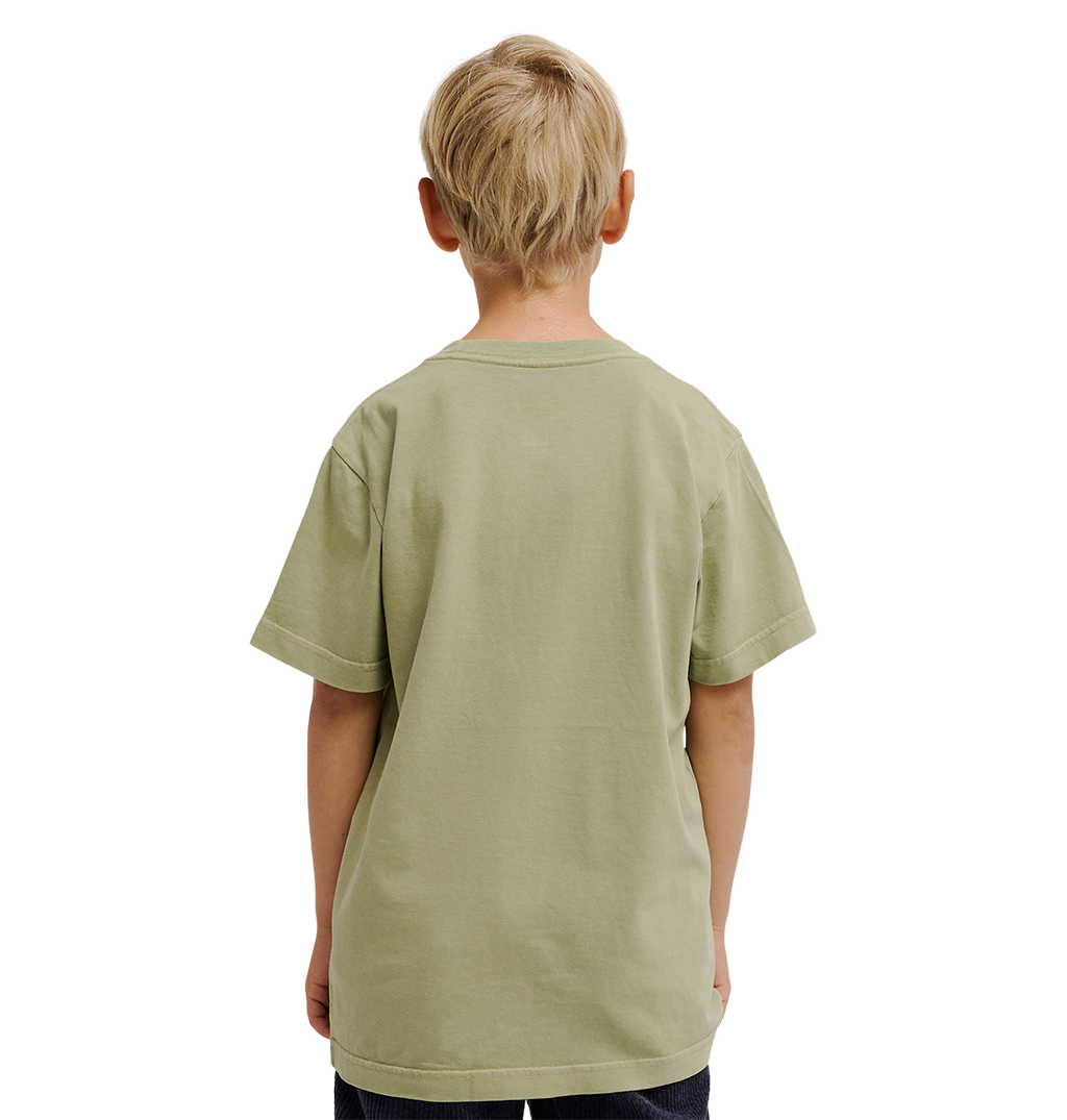A young boy with short blond hair stands facing away, wearing the DC Shoes DC Speratic Tee Youth in a relaxed fit pale green graphic style and dark pants against a plain white background.