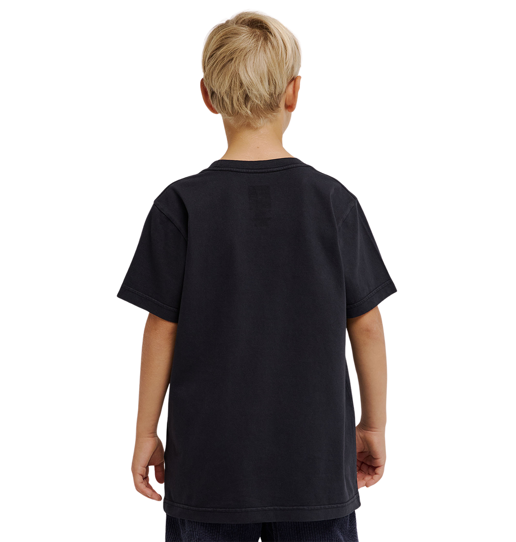 A young boy with short blond hair is standing with his back to the camera, wearing a loose-fitting, plain black DC Speratic Tee Youth by DC Shoes and dark shorts against a light gray background.