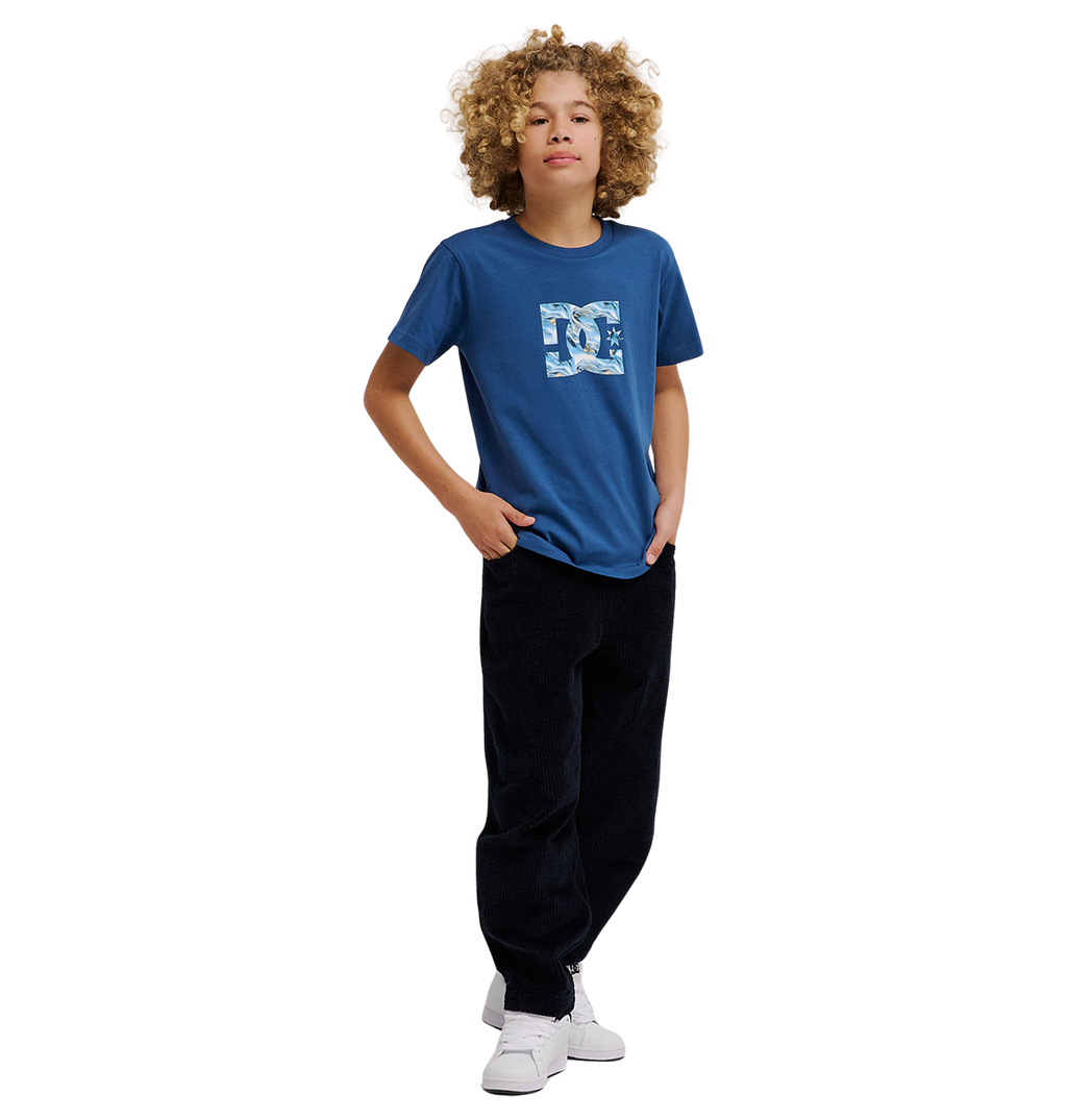 A young person with curly blond hair wears the DC Shoes DC Star Filled Tee Youth—a relaxed-fit blue graphic skate t-shirt with bold star artwork—paired with black pants and white sneakers, standing confidently with one hand in their pocket against a plain white background.