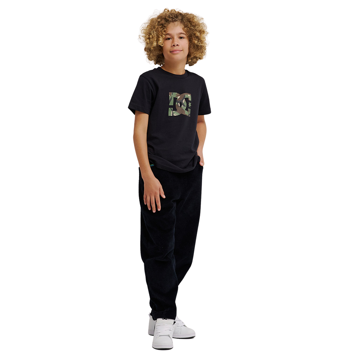 A young person with curly hair stands against a white background, wearing the DC Shoes DC Star Filled Tee Youth—a black graphic skate t-shirt featuring signature DC star artwork—paired with relaxed-fit black pants and white sneakers, one hand in their pocket.