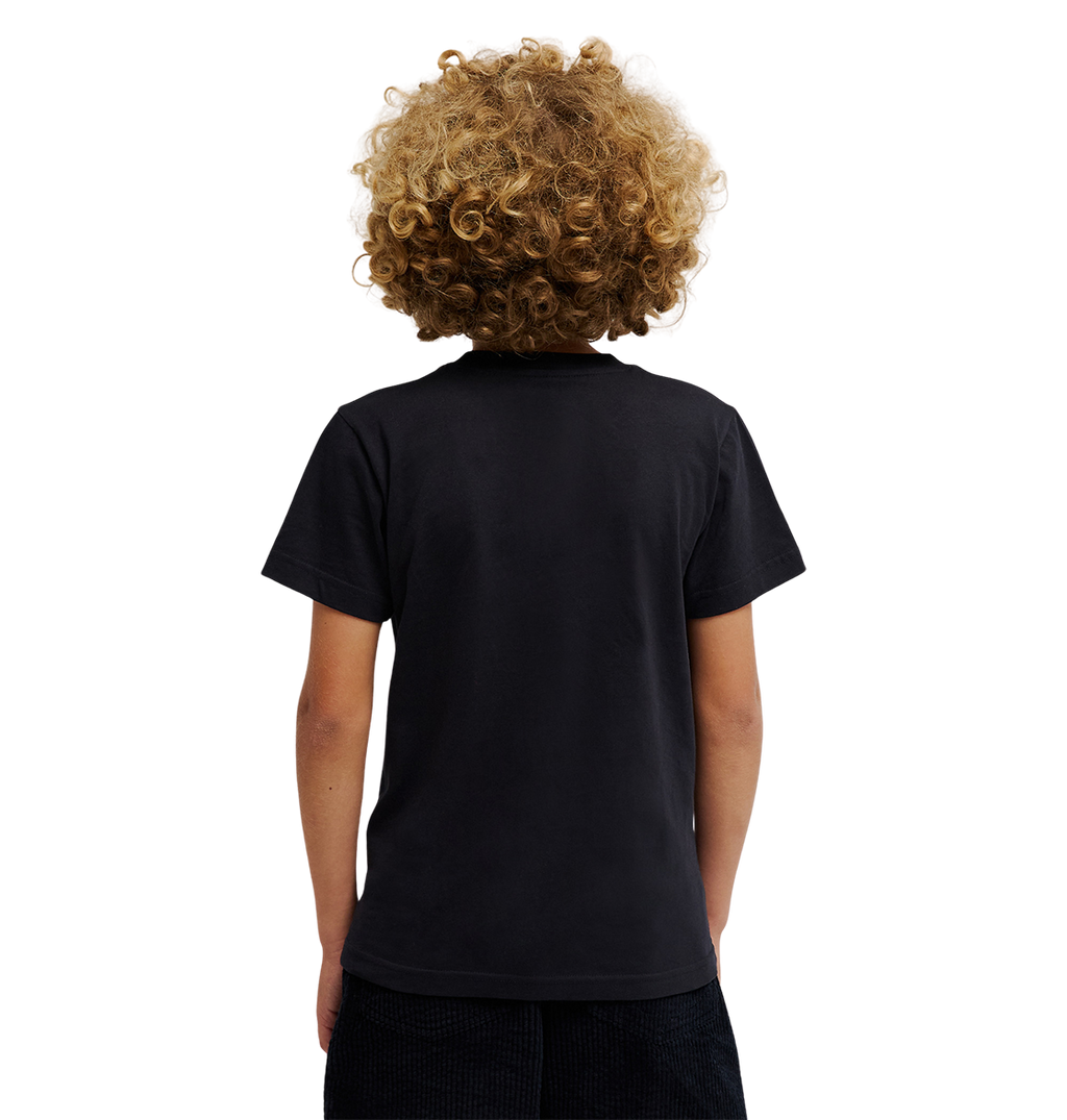 A child with curly blond hair, dressed in a relaxed fit DC Shoes Side Flame Tee Youth featuring subtle flame graphics, and dark shorts, stands with their back to the camera against a light, plain background.