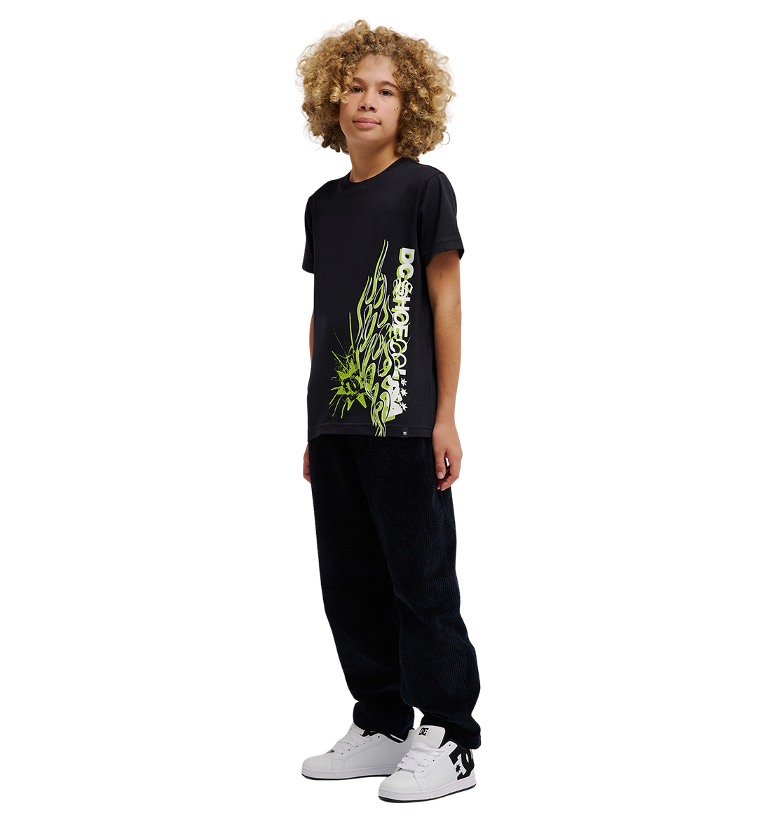 A young person with curly blond hair stands facing forward, wearing a relaxed fit DC Shoes Side Flame Tee Youth in black, featuring green flame graphics, paired with black pants and white sneakers. The background is plain white.