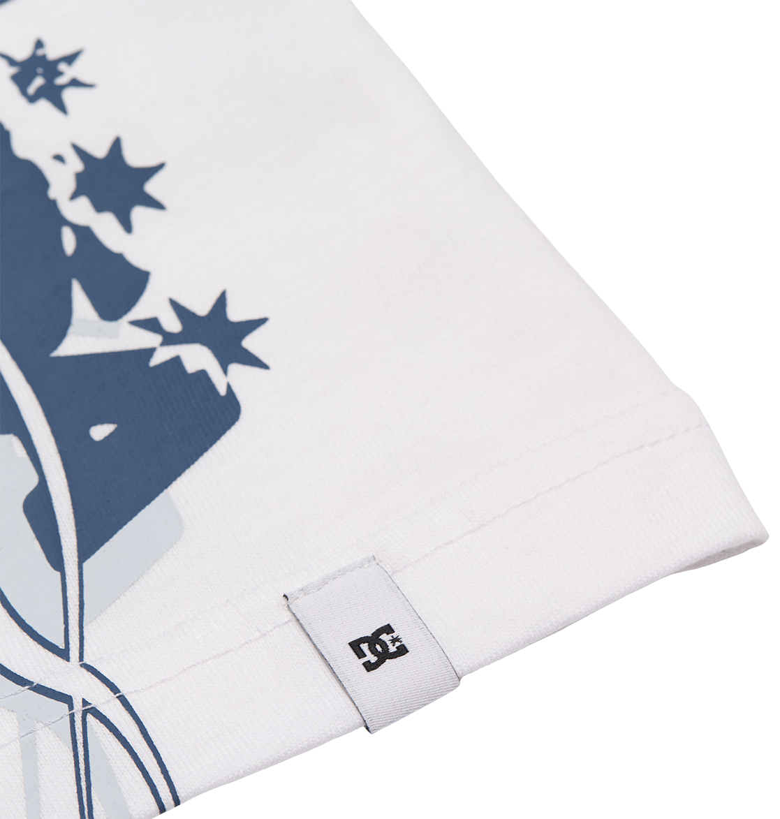 Close-up of the hem on the DC Shoes Side Flame Tee Youth in white, featuring a small DC logo tag. A section of the tee’s bold blue graphic with stars is visible, reflecting its relaxed fit and skate-inspired style.