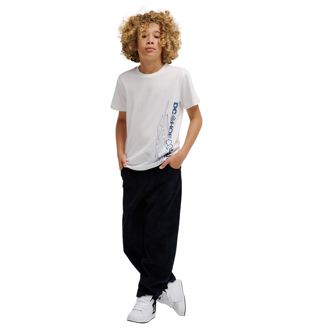 A boy with curly hair wears the DC Shoes DC Side Flame Tee Youth, a white graphic skate tee featuring bold flame artwork, paired with black relaxed fit pants and white sneakers. He stands with his hands in his pockets against a plain white background.