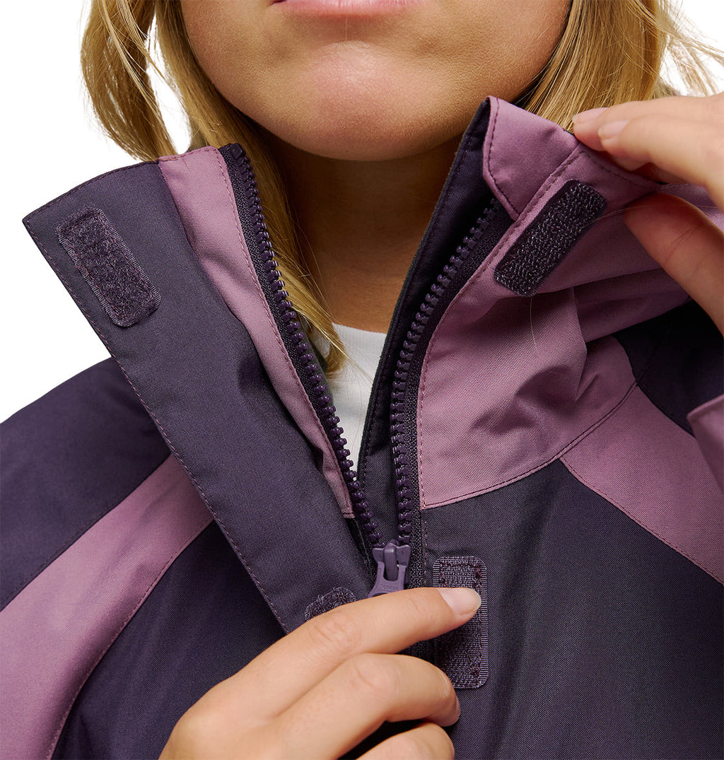 A person zips up the collar of the DC Shoes Everfrost Jacket, highlighting its two-tone purple insulated design and Velcro closures, with a focus on their hands and the jacket’s detailing.