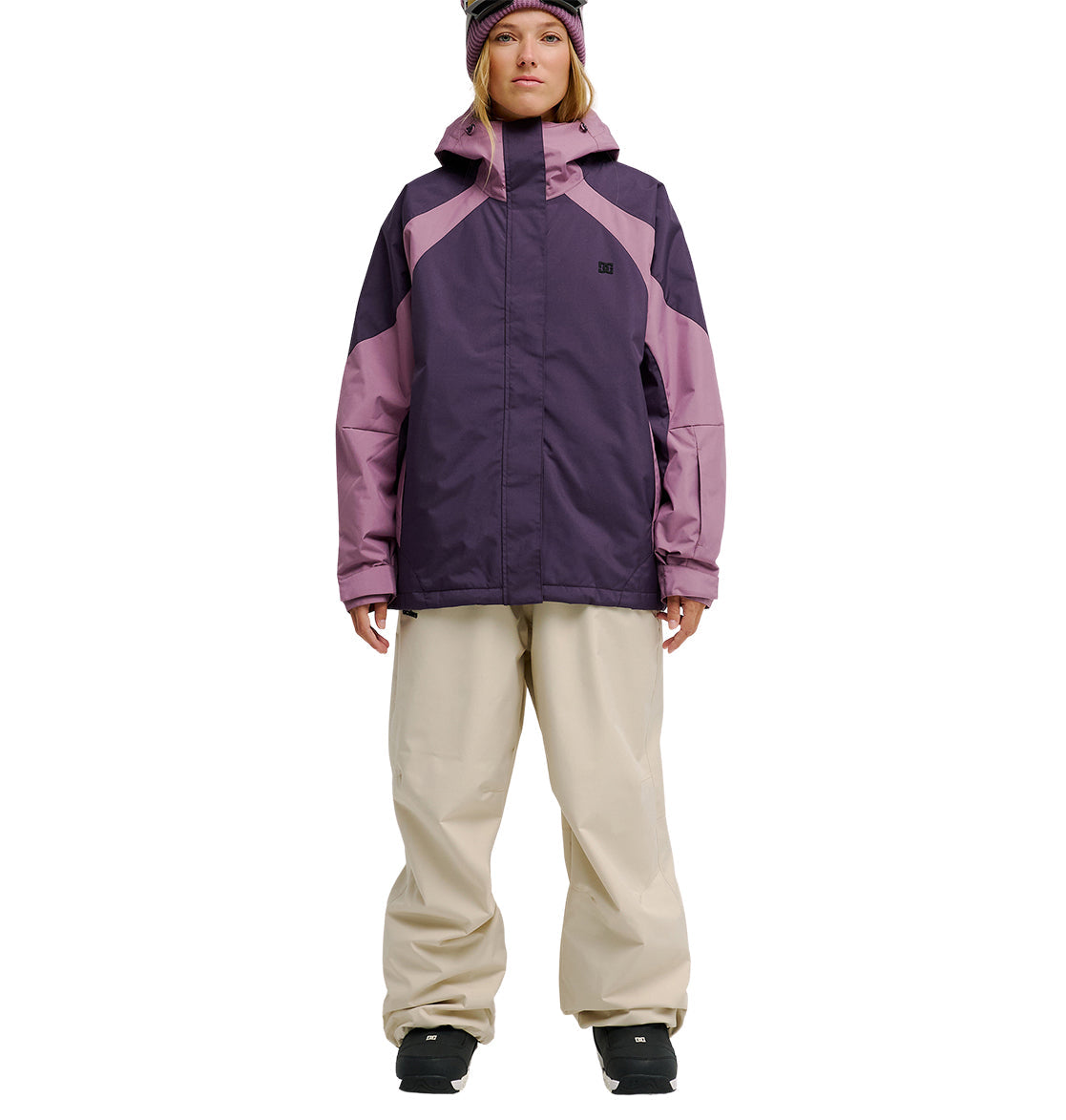 A person wearing the DC Shoes Everfrost Jacket—a purple and pink waterproof jacket—along with beige snow pants, black boots, and a purple hat stands facing forward against a white background, ready for peak snow performance.