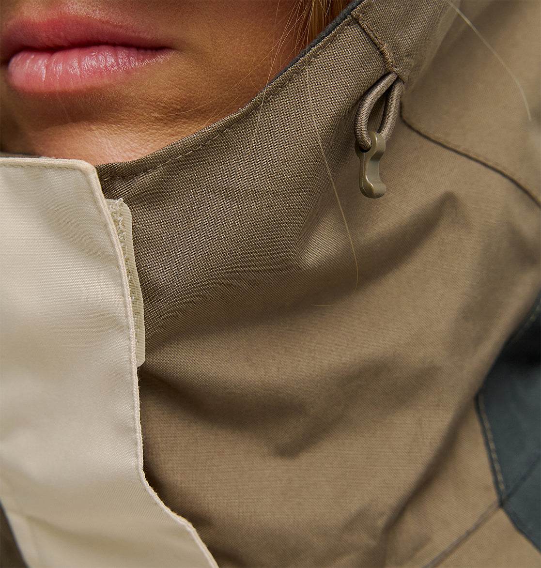 Close-up of a person’s lower face, showing pink lips and part of the chin. The person is wearing the DC Shoes Everfrost Jacket—an insulated outerwear piece in tan and beige, featuring a high collar with a visible drawstring.