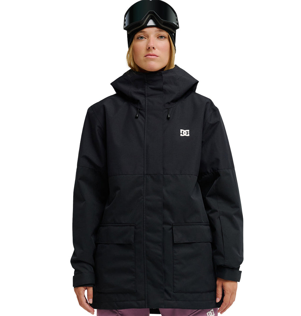 A person wearing a black DC Shoes Cruiser Jacket—an insulated, fully waterproof snow jacket—paired with pink pants, black ski goggles, and a black beanie stands facing forward against a white background.