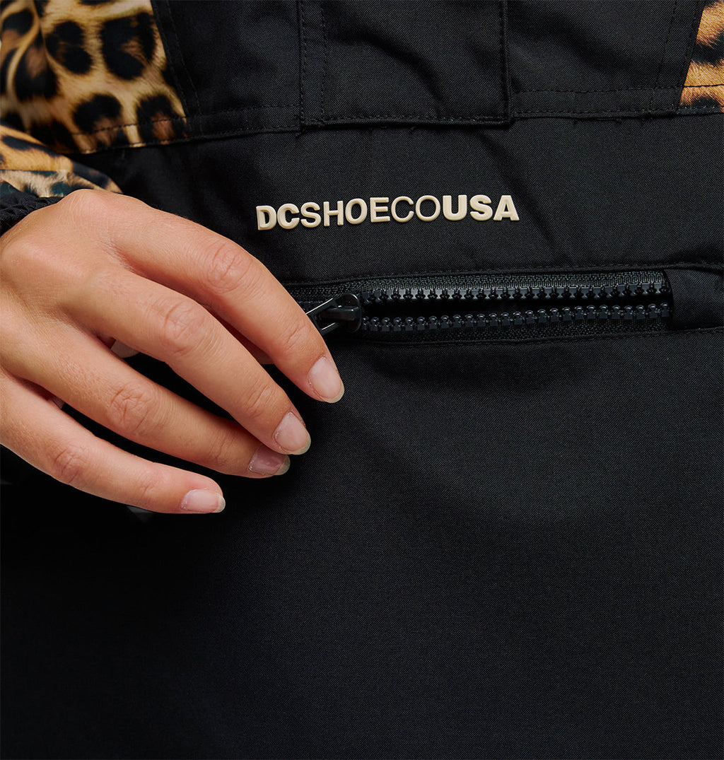 A hand with neatly manicured nails pulls the zipper of a black Chalet Anorak jacket by DC Shoes, featuring the DCSHOECOUSA logo and a leopard print detail on the sleeve, designed for breathable protection.