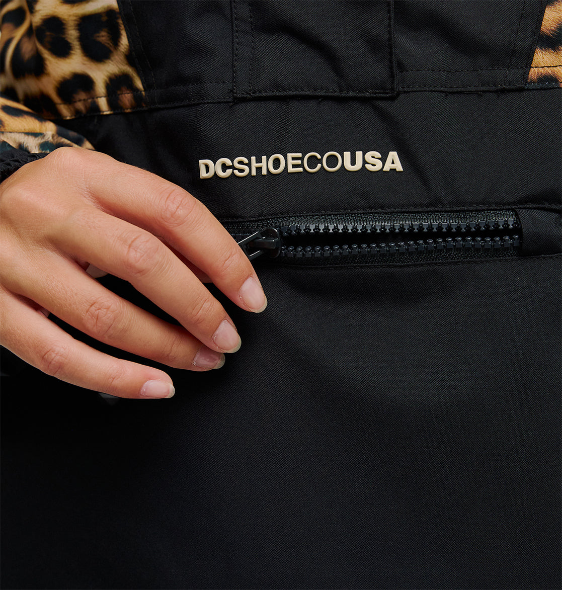 A hand with neatly manicured nails pulls the zipper of a black Chalet Anorak jacket by DC Shoes, featuring the DCSHOECOUSA logo and a leopard print detail on the sleeve, designed for breathable protection.