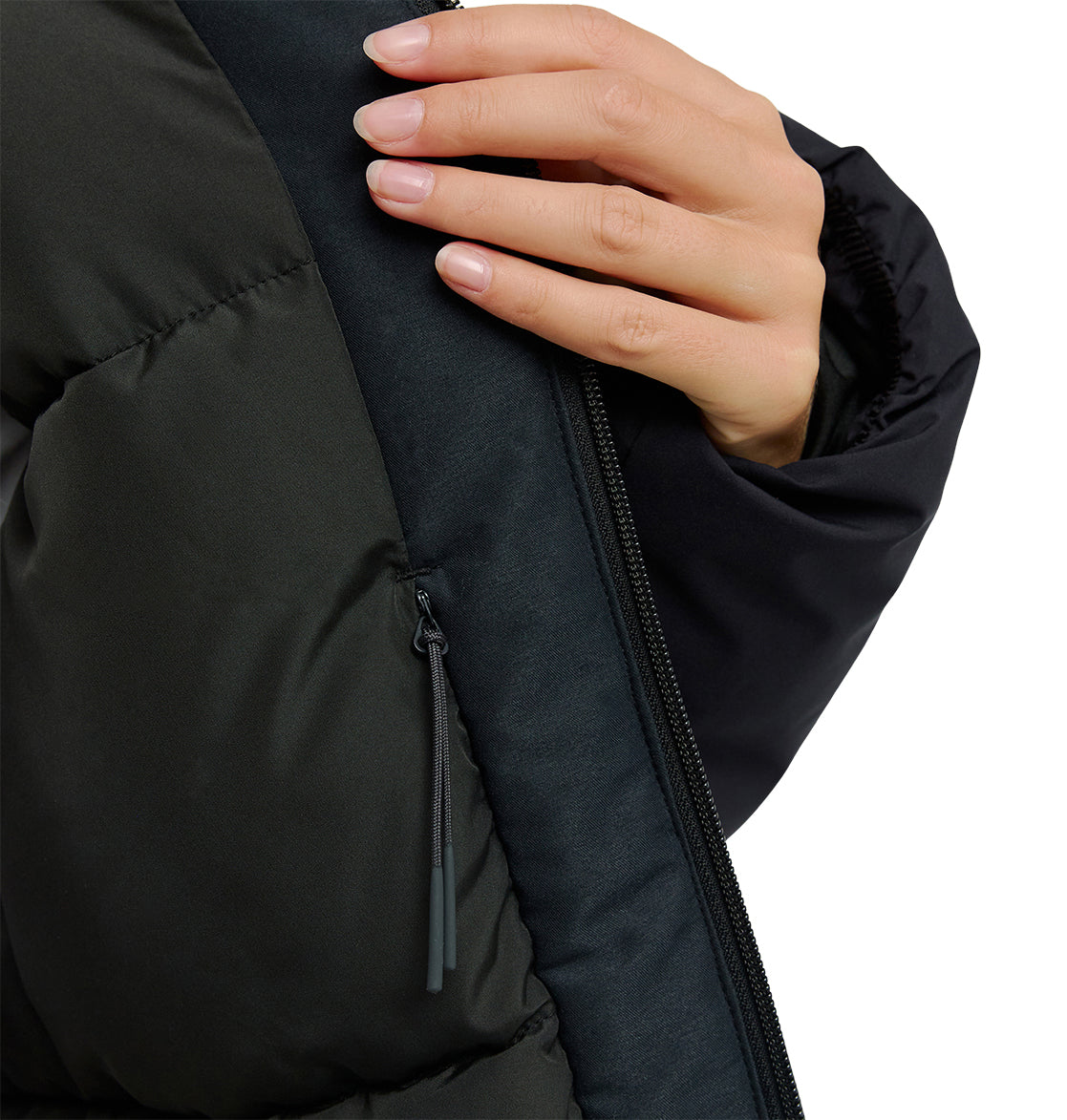 A hand with neatly manicured nails holds the zipper of the DC Shoes Aura Puffer Jacket, highlighting its waterproof black fabric and a small zippered pocket on the chest.