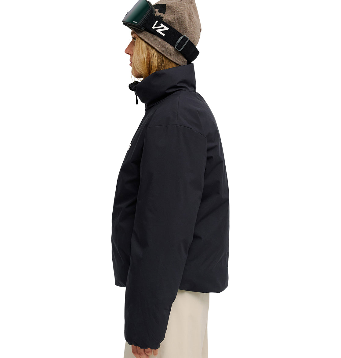 Wearing the DC Shoes Aura Puffer Jacket, tan pants, a tan beanie, and ski goggles, a person stands in profile facing left against a white background.
