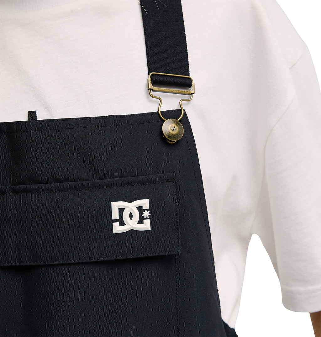 A person wears the Valiant Bib Pants by DC Shoes—black overalls with a front pocket, gold buckle, and a signature logo featuring interlocking DC letters with a star—paired with a white T-shirt. Designed for breathable protection and comfort during everyday wear.