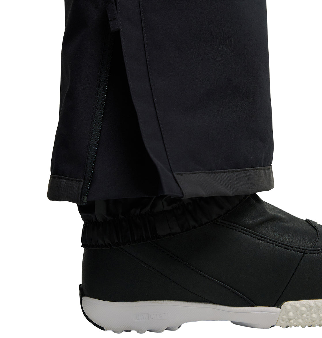 Close-up of a person wearing DC Shoes Valiant Bib Pants in black, featuring a zipper near the ankle for breathable protection and covering the top of a black shoe with a white sole and textured tread, set against a white background.