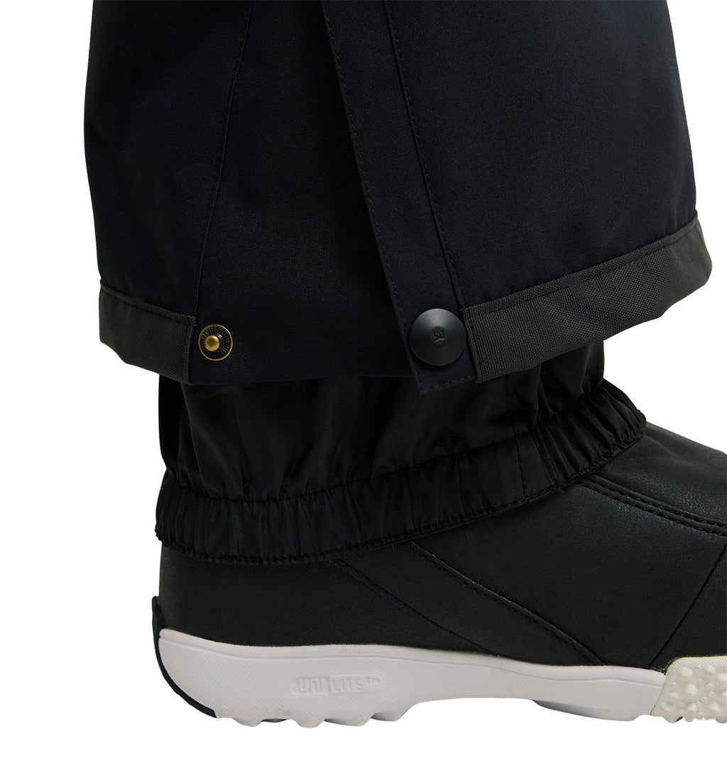 Close-up of a person wearing the DC Shoes Nonchalant Pant in black—an insulated style featuring elastic cuffs over black shoes. These pants offer button and snap closures at the ankle, blending snow-ready performance with added waterproof protection for cold-weather comfort.
