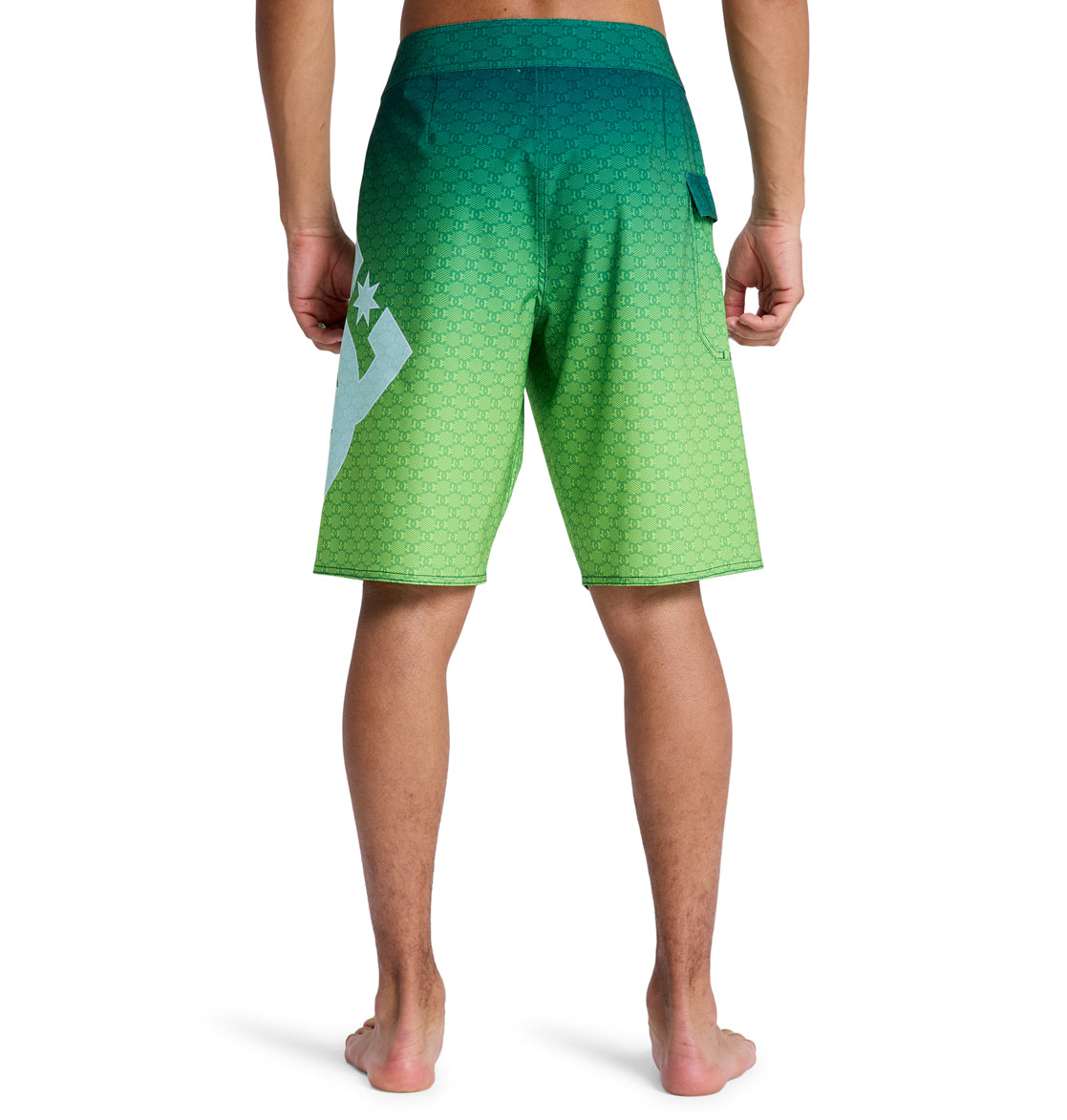 A person is seen from the back, barefoot, wearing Reebok Lanai 21 swim trunks featuring a green gradient with a subtle hexagonal pattern and a bold white graphic on the left side.
