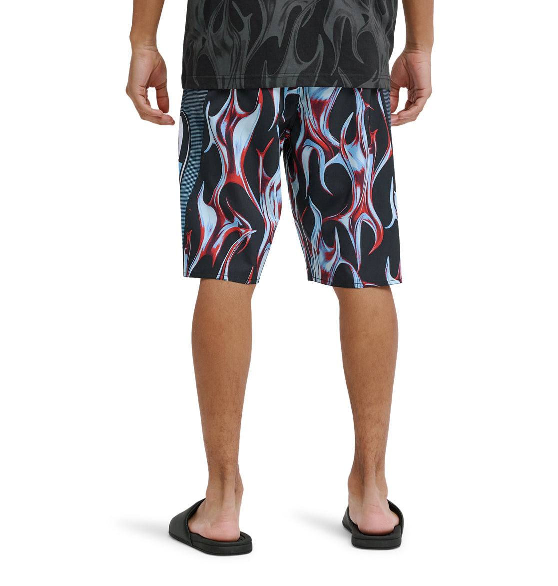 A person stands facing away, wearing Reebok Sylem 21 black and red flame-patterned shorts, a dark shirt, and black slide sandals, shown from the knees down against a white background.