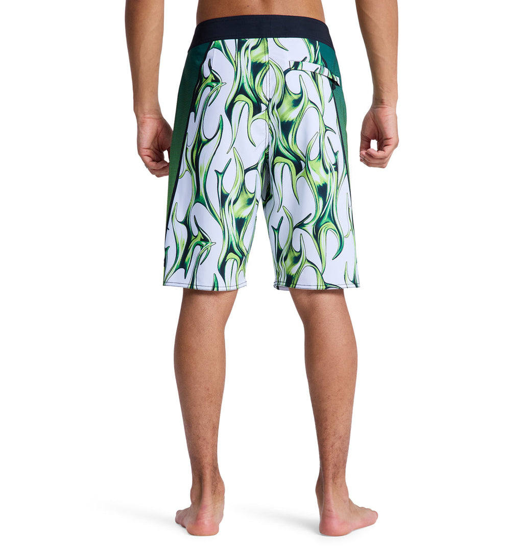 A person stands barefoot, facing away, wearing Reebok Sylem 21 board shorts with a black waistband and side panels, featuring a bold white and green patterned design with wavy green flames.