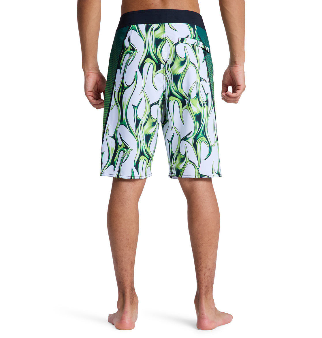 A person stands barefoot, facing away, wearing Reebok Sylem 21 board shorts with a black waistband and side panels, featuring a bold white and green patterned design with wavy green flames.