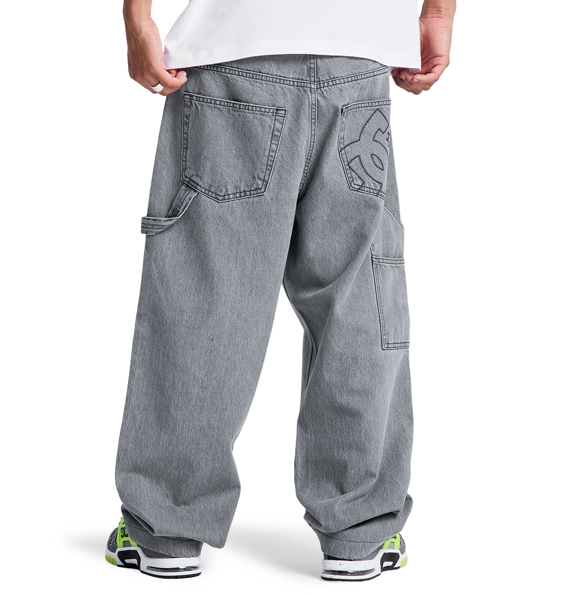 A person stands facing away, wearing DC Shoes Men's Baggy Denim Carpenter Pant with large pockets and a white graphic on the back pocket, styled with a white shirt and chunky sneakers featuring neon green accents.