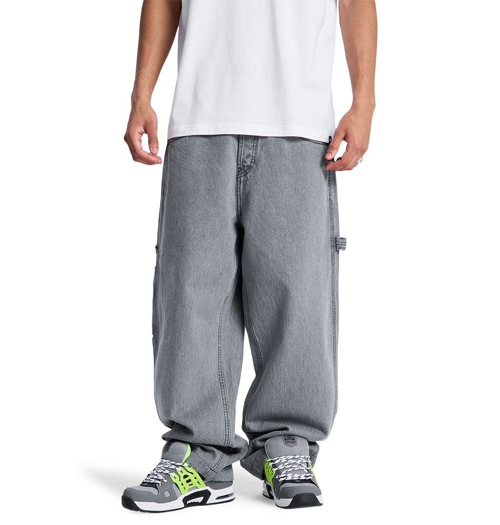 A person stands facing forward, hands relaxed, wearing a white T-shirt and DC Shoes Men's Baggy Denim Carpenter Pant in gray, paired with gray sneakers featuring green and black accents. Photo is cropped just below the shoulders.