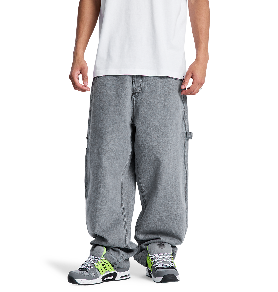 A person stands facing forward, hands relaxed, wearing a white T-shirt and DC Shoes Men's Baggy Denim Carpenter Pant in gray, paired with gray sneakers featuring green and black accents. Photo is cropped just below the shoulders.