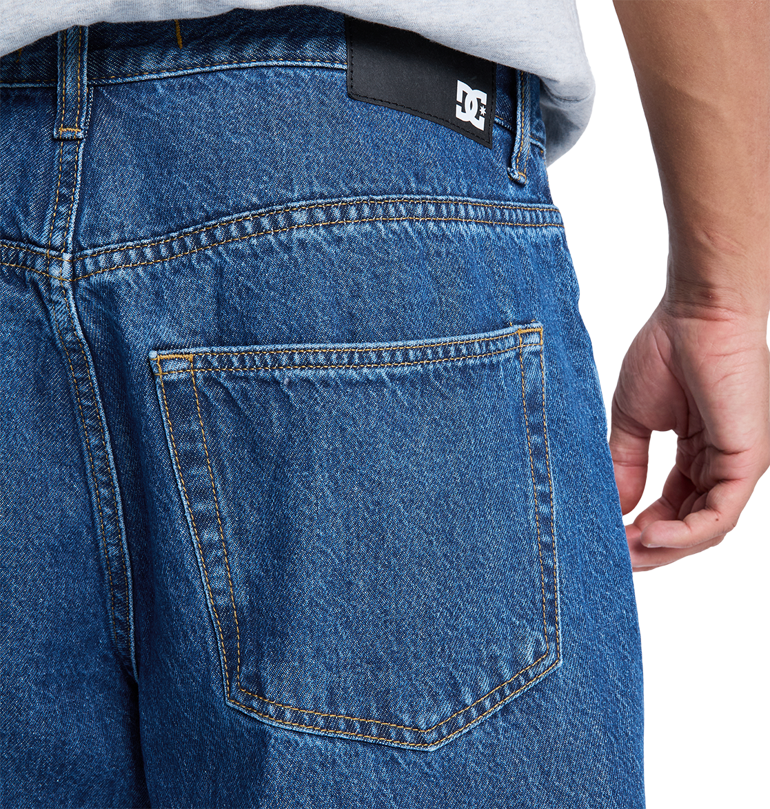 Close-up of the back pocket on DC Shoes Men's Baggy Denim Pant in blue heavyweight denim, showing visible stitching and a black patch with the white DC logo near the waistband. A person's hand rests by their side.