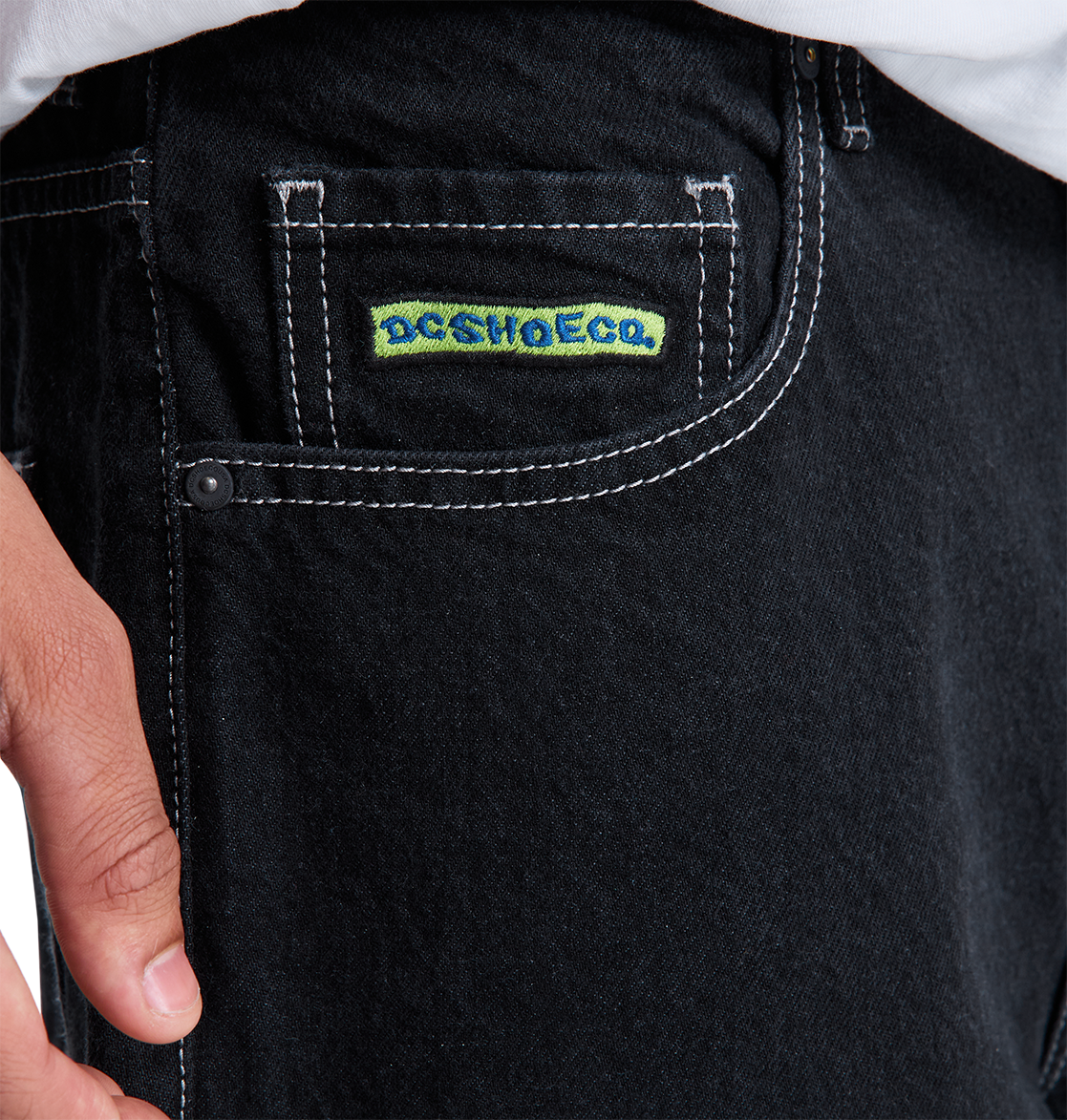 Close-up of a person’s hand near the pocket of DC Shoes Men's Baggy Denim Pant, featuring white stitching and a small DCSHOECO label. They are wearing a white shirt tucked into the heavyweight denim jeans.