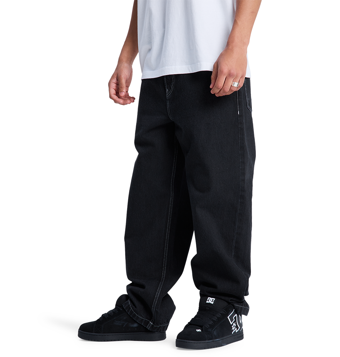 A model wears DC Shoes Men's Baggy Denim Pant and black DC skate shoes with a visible logo, paired with a white T-shirt. Cropped at the neck, the image highlights the relaxed, lower-body skate style against a white background.