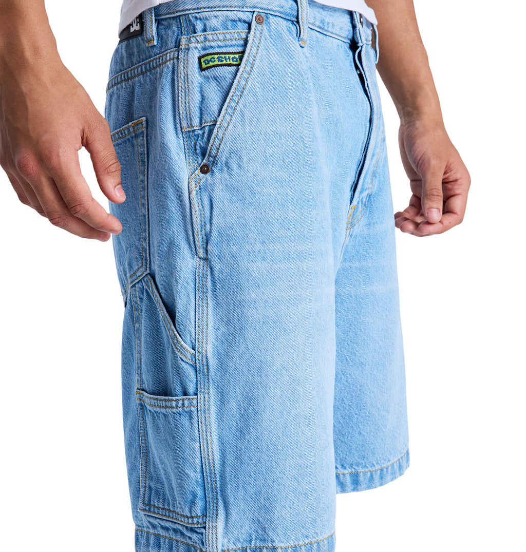 A person wears Reebok Baggy Denim Carpenter Shorts in light blue with side pockets and a small green patch above one pocket; only the lower torso and hands are shown.