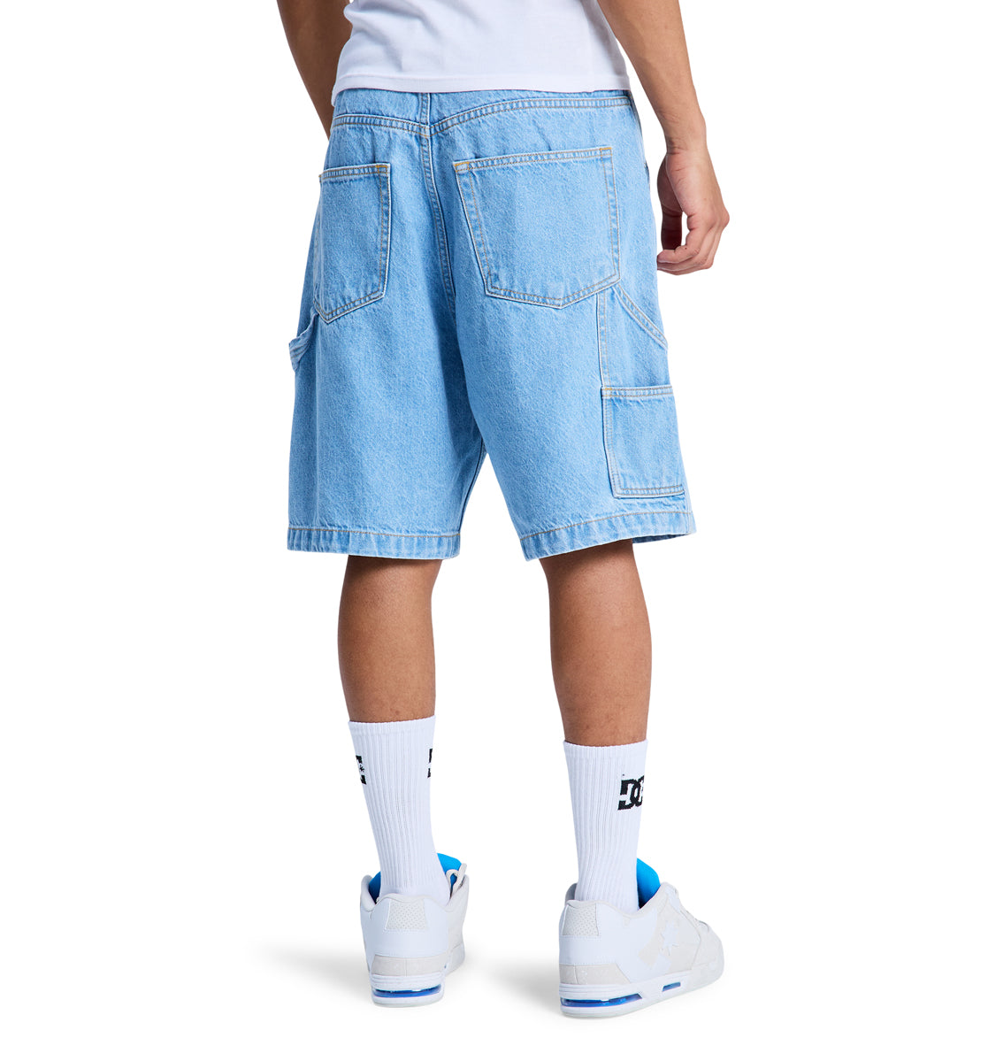 A person wearing Reebok Baggy Denim Carpenter Shorts, featuring large back pockets, paired with white DC socks and white sneakers, is shown from the back against a plain white background.