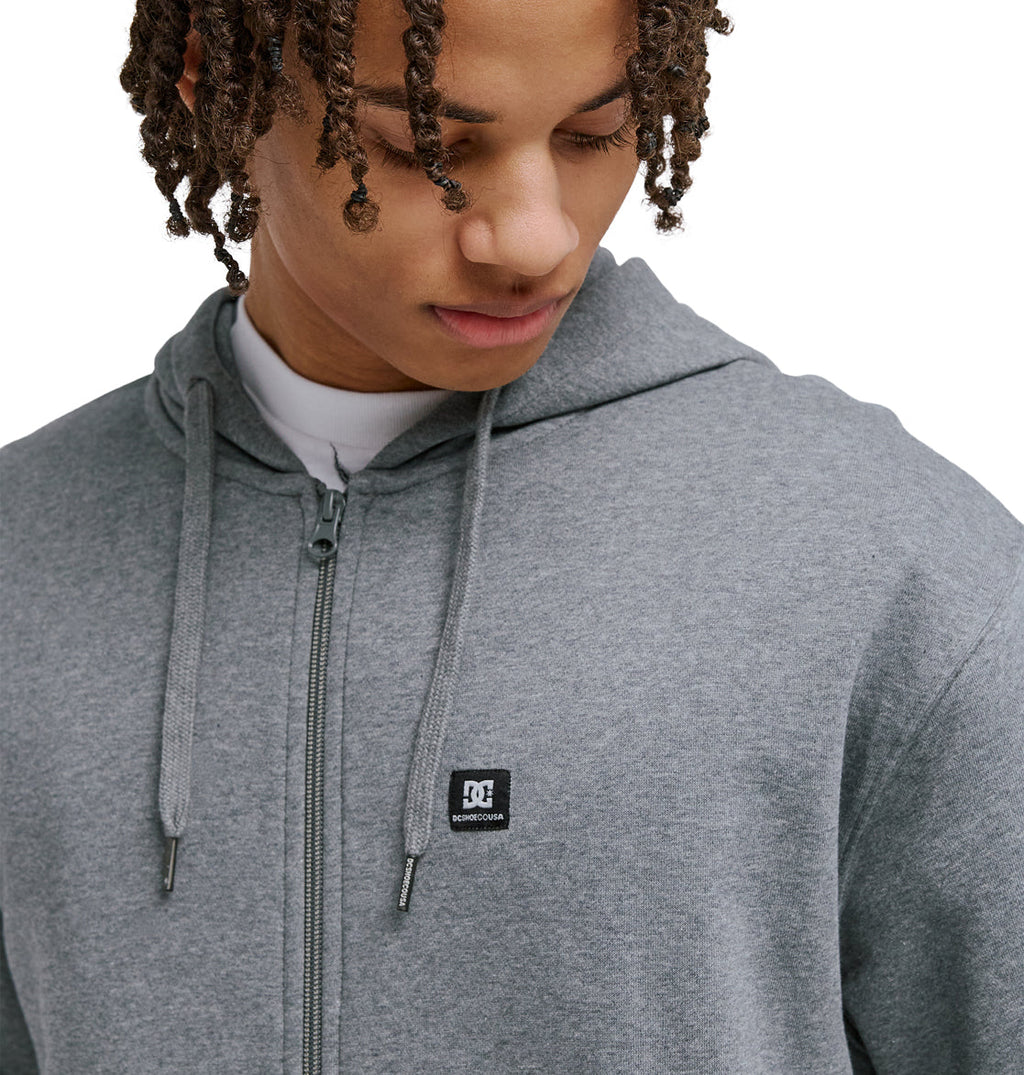 A person with curly hair, wearing the Reebok Lorion Zh gray zip-up hoodie featuring a small black and white logo on the chest, looks down against a plain white background.