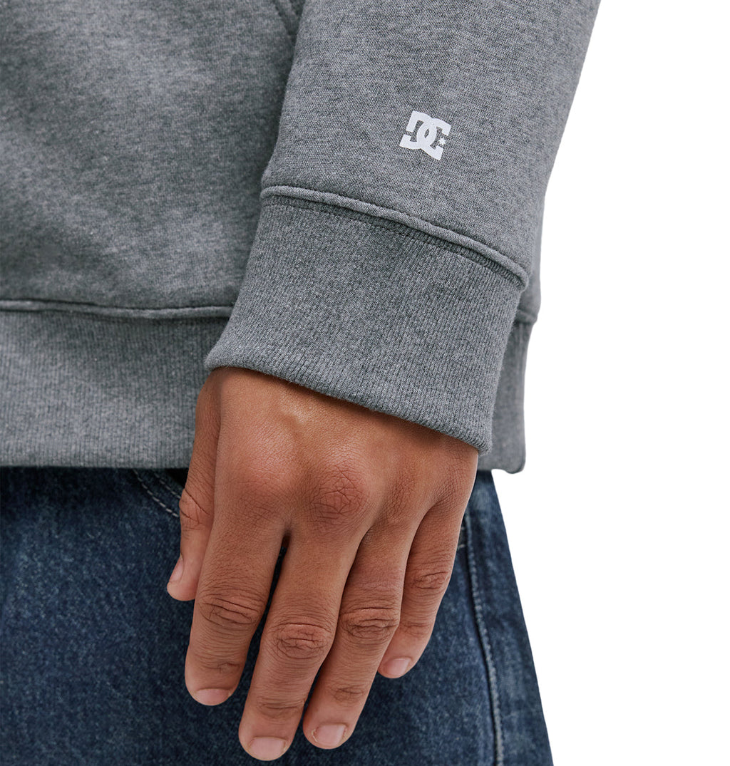 A close-up of a person’s hand resting at their side, wearing the Reebok Lorion Zh grey sweatshirt with a small white logo on the sleeve and blue denim jeans.