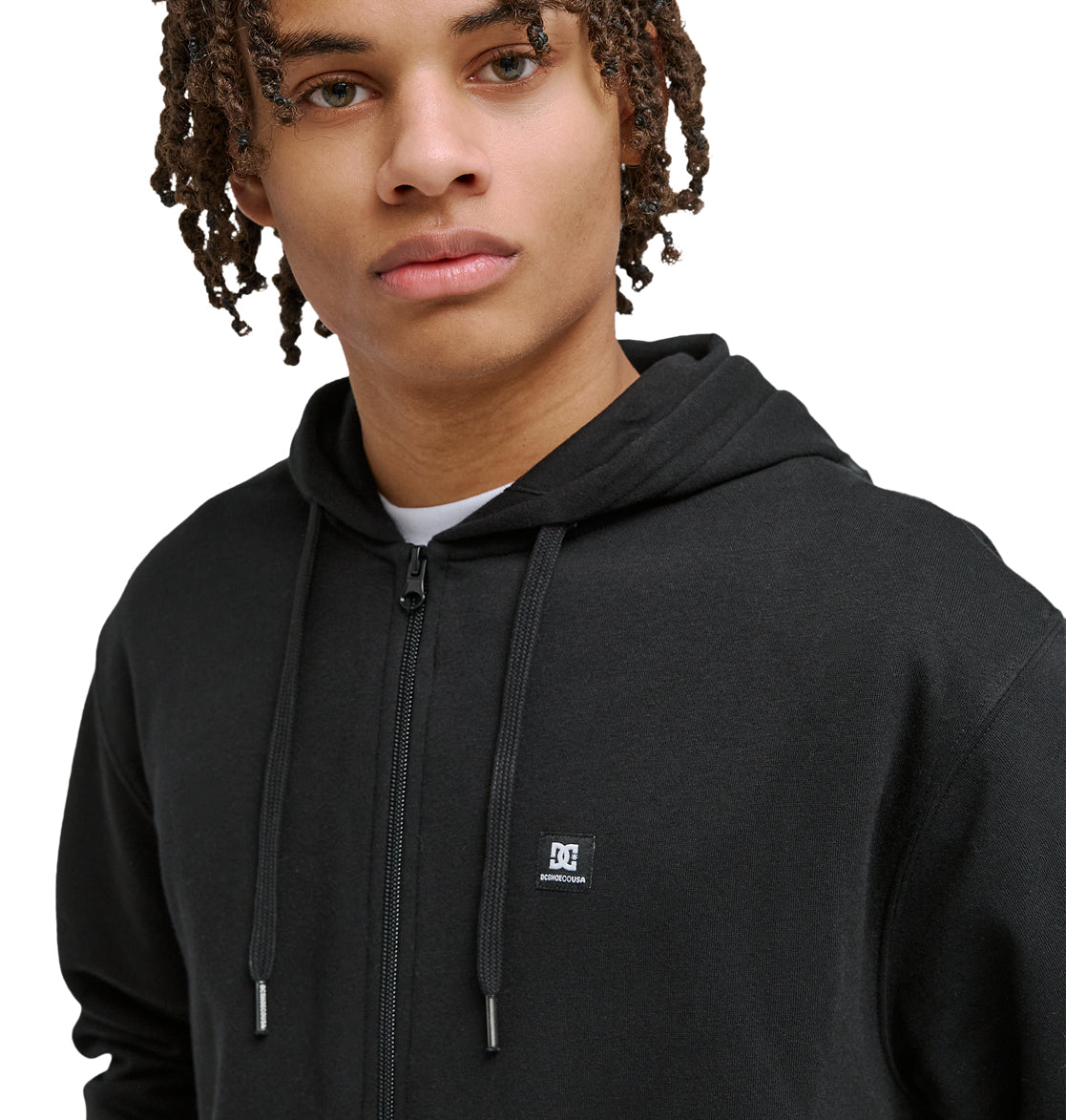 A young person with medium brown skin and curly hair wears the Reebok Lorion Zh black zip-up hoodie, featuring a small white chest logo, while looking slightly past the camera against a plain white background.