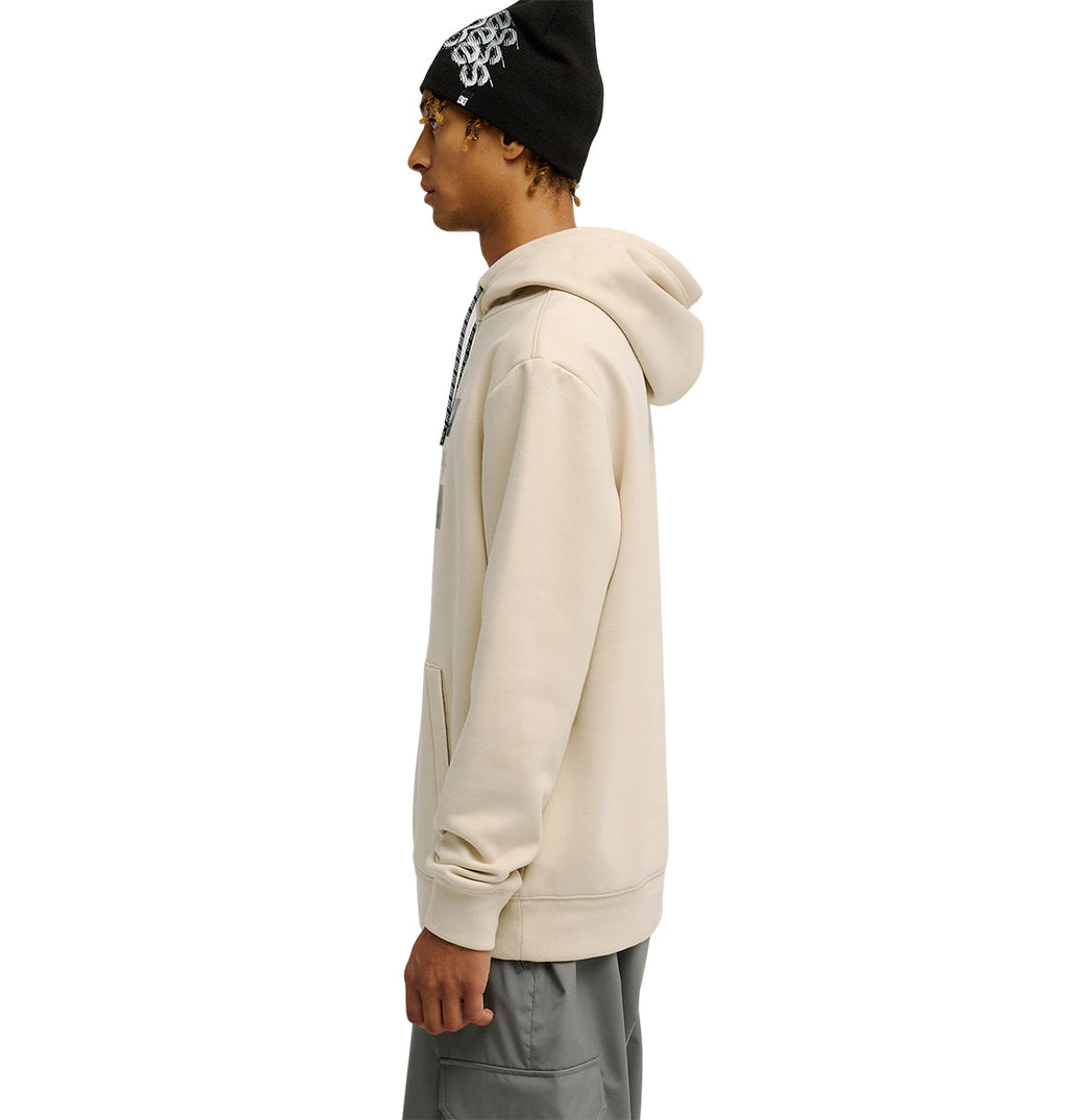 Wearing the DC Shoes Snowstar hoodie in beige, paired with gray pants and a black beanie featuring a white design, a person stands in profile against a white background, highlighting the comfortable fit and weather protection this outfit offers.