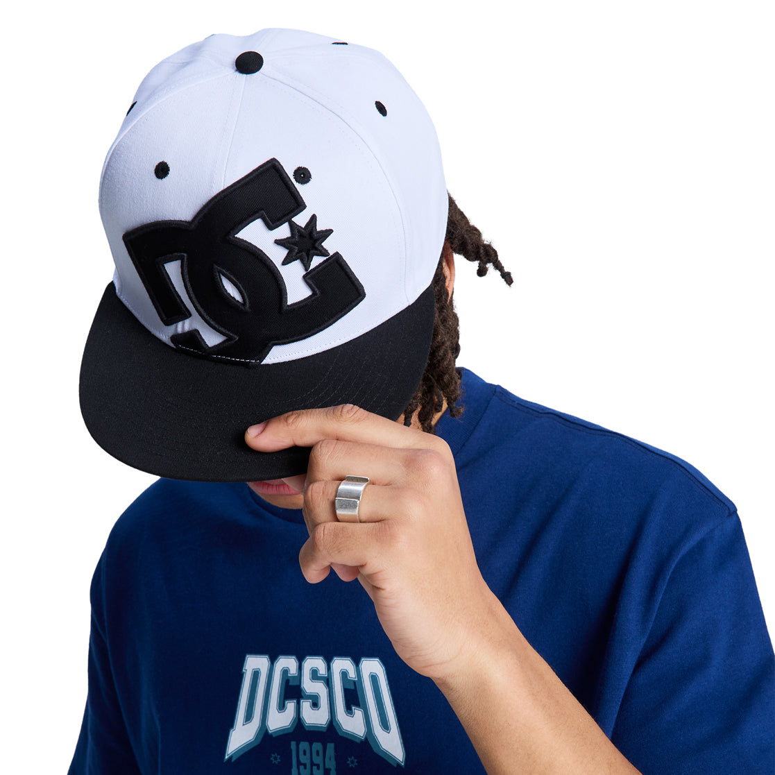 A person in a blue DCSCO 1994 shirt and a Reebok DC Double That Cap tilts the brim to partially cover their face, revealing a medium skin tone and a silver ring on one finger.