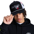A person wearing a black hoodie and a Reebok Side Star Trucker hat with embroidered designs lifts the hat's brim, looking directly at the camera against a white background.
