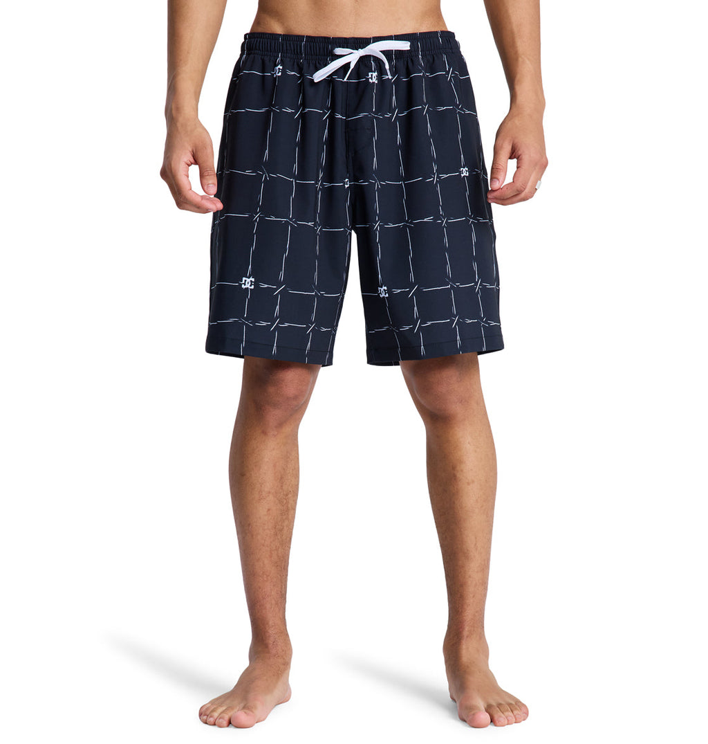 A person stands barefoot, shown from waist to feet in Reebok DC Wired Volley 19 black swim trunks with a white grid pattern and white drawstring, against a plain white background.