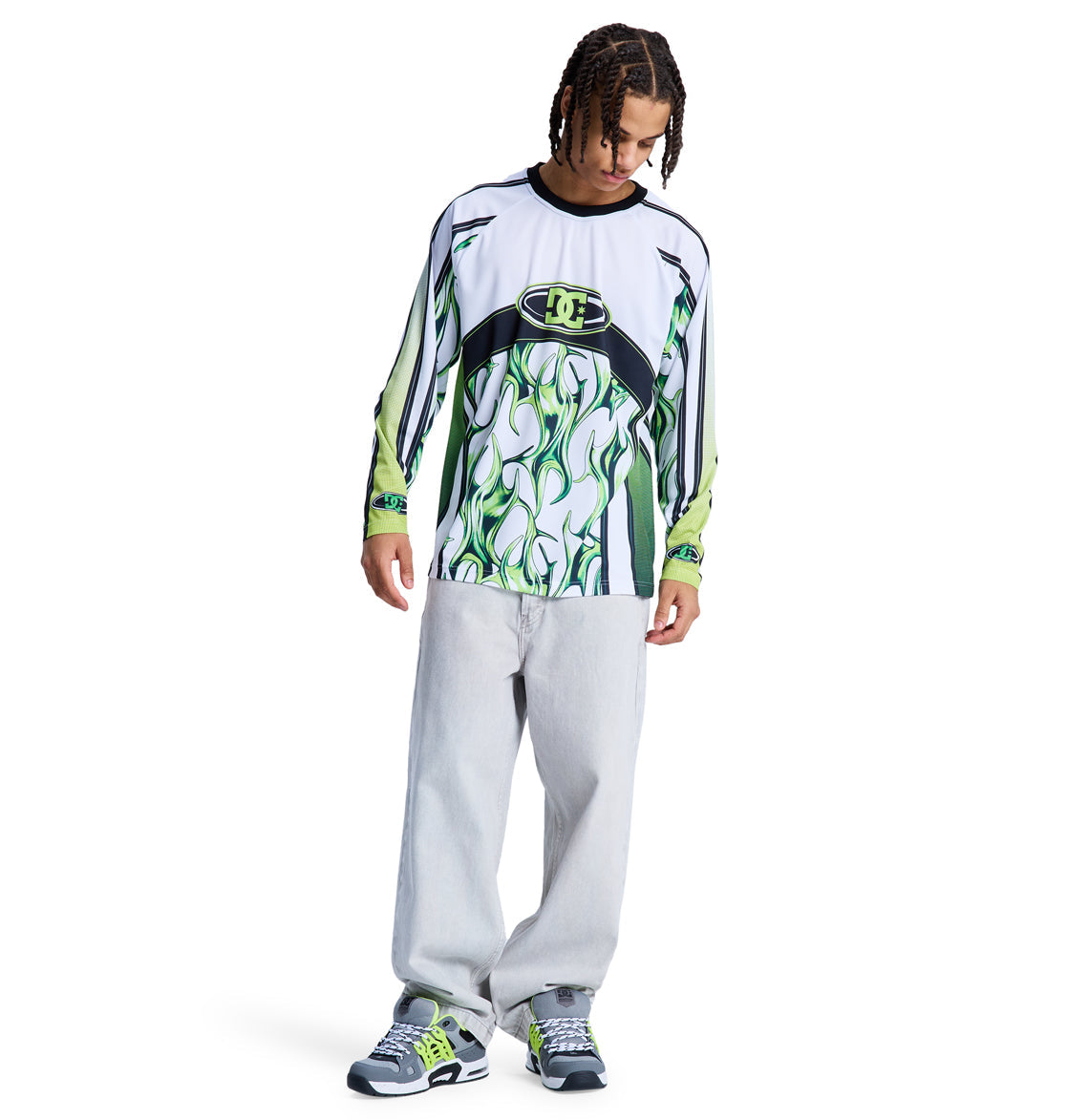 A young man with braided hair wears the Reebok Sylem 2 Ls long-sleeve shirt featuring abstract green and white patterns, paired with loose light gray jeans and gray sneakers with green accents, standing against a white background.