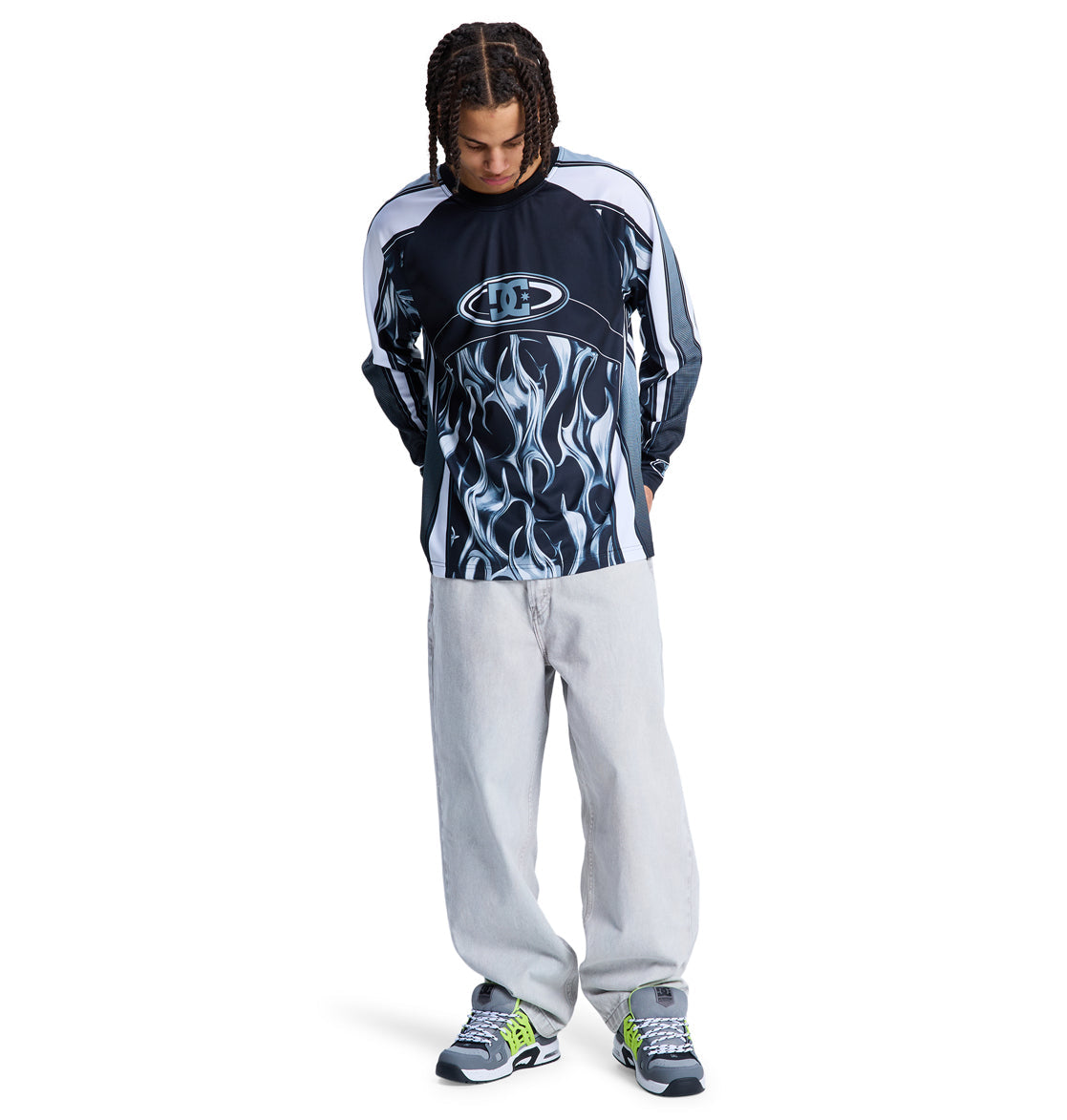 A person with braided hair wearing the Reebok Sylem 2 Ls black and white flame-print long-sleeve shirt, light gray baggy jeans, and green-accented sneakers stands with hands in pockets against a white background.