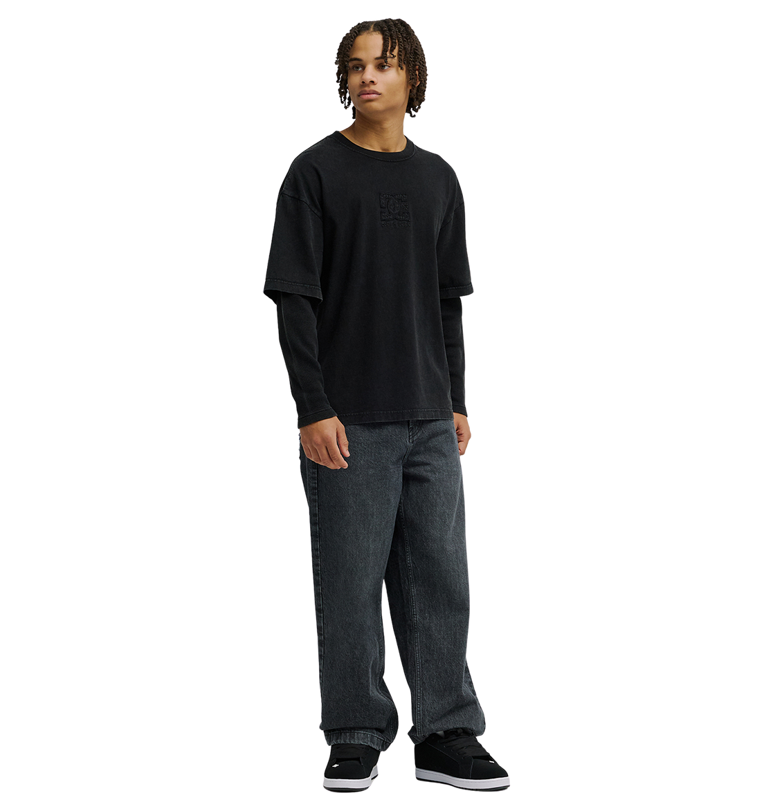 A person with medium-length curly hair wears the DC Shoes Men’s DC Twofer Long Sleeve under a black T-shirt, paired with loose dark jeans and black sneakers, embracing a streetwear style while standing against a plain white background.