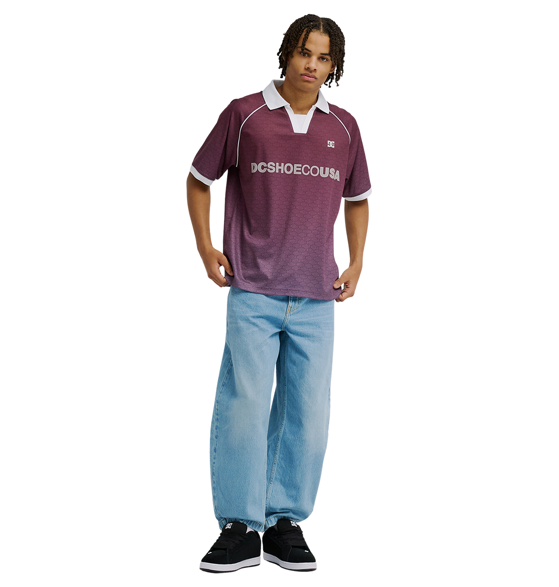 A person stands against a plain white background wearing the Men's DC Soccer Top by DC Shoes in maroon, paired with light blue baggy jeans and black sneakers.